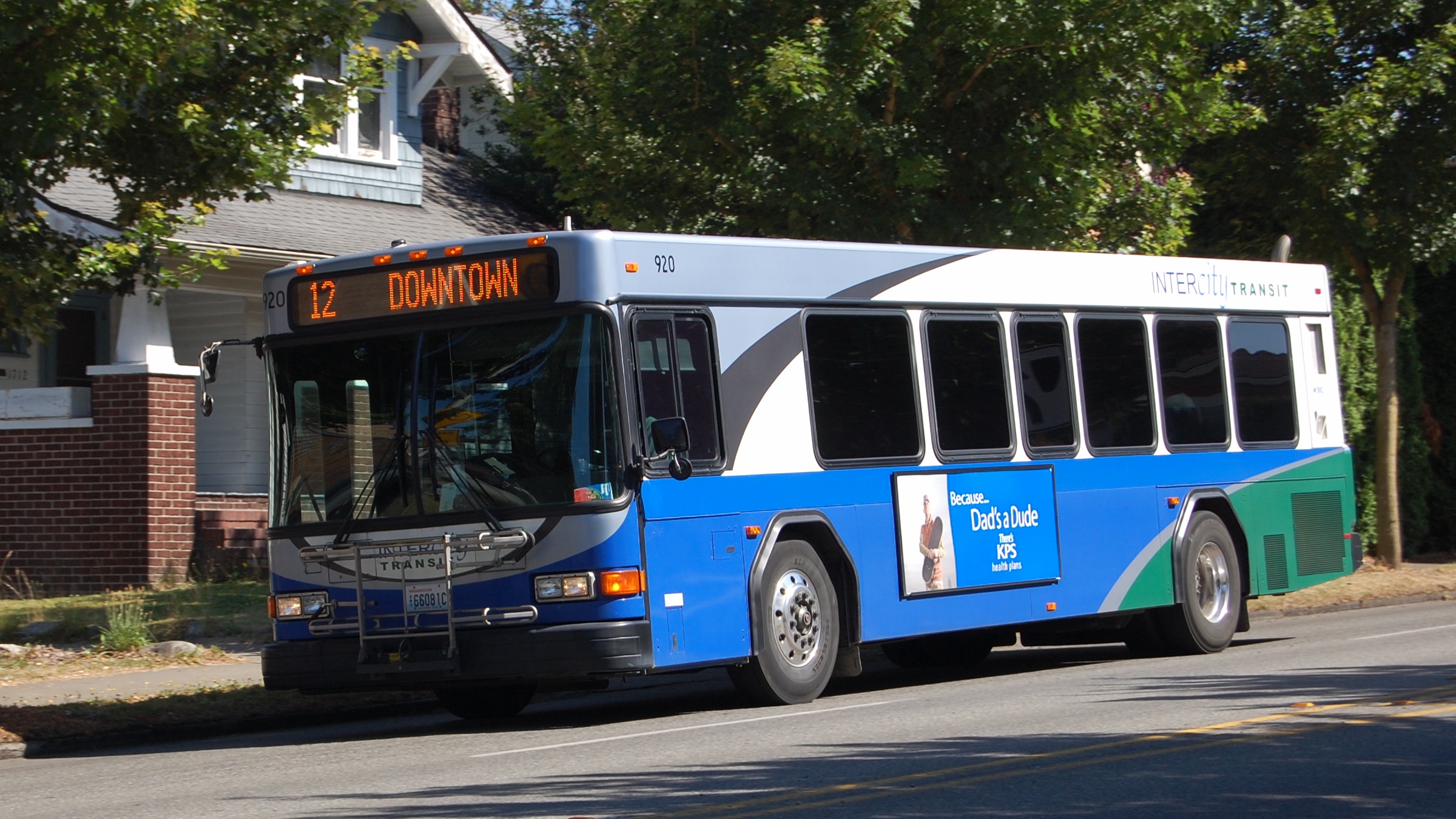 A blue and teal Intercity Transit bus on Route 12 heading downtown on a tree-lined street.