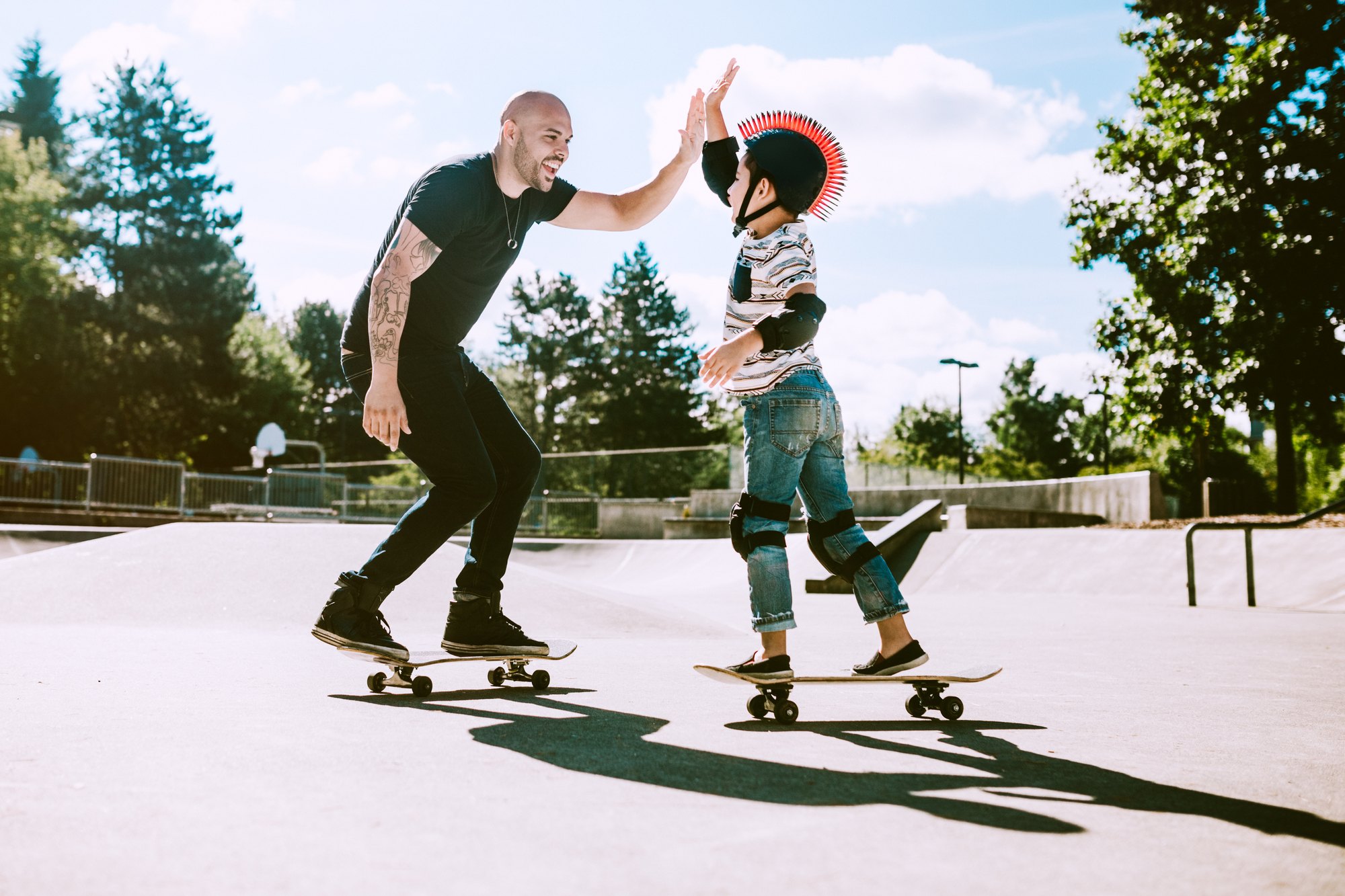 Adult and child on skateboards giving a high-five at a sunny outdoor skate park.
