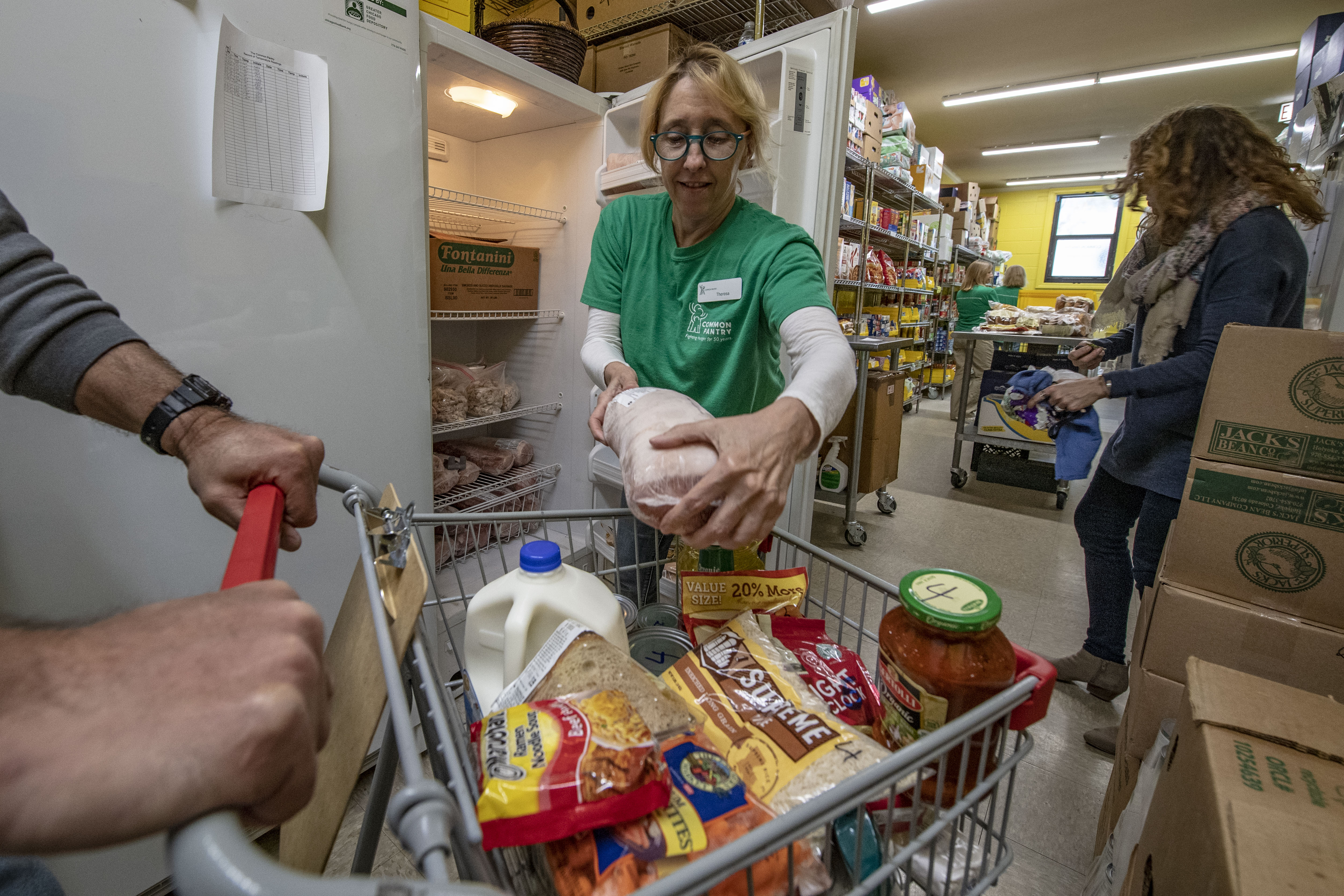 Volunteer in green shirt handing food to a person pushing a grocery-filled cart.
