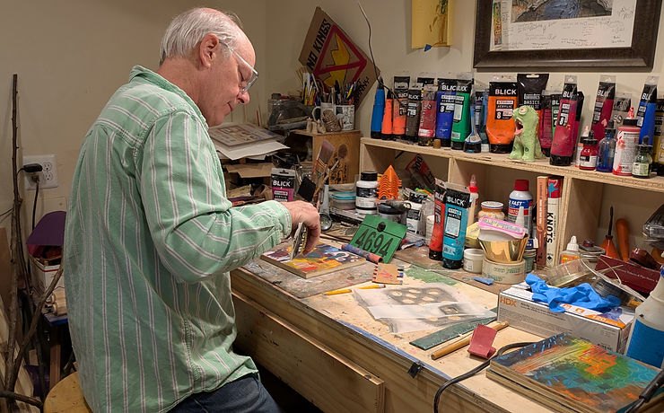 A man in a green striped shirt and glasses works at a cluttered art studio desk with paint tubes on shelves behind him.
