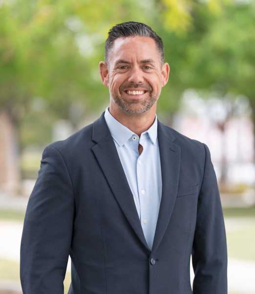 Smiling man in a navy blazer standing outdoors in front of blurred trees.