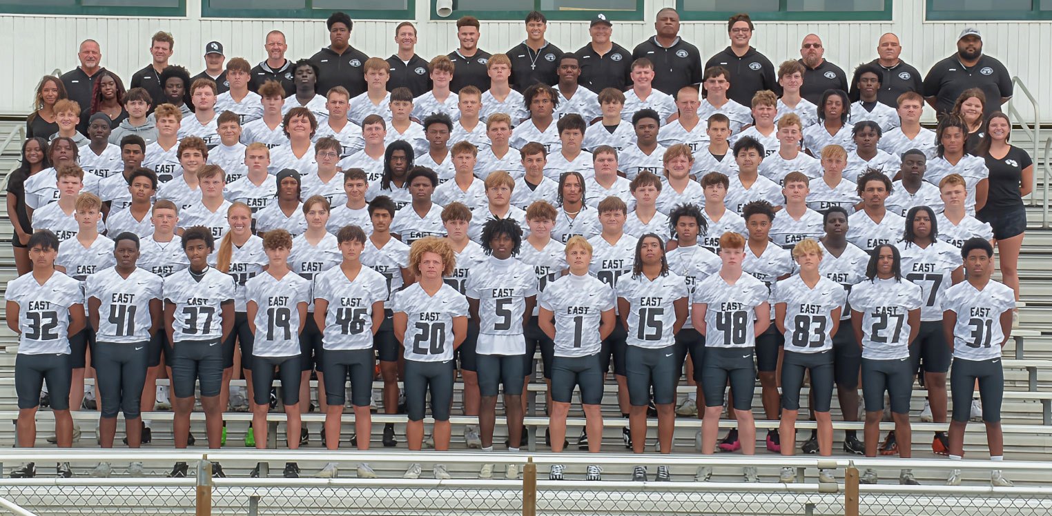 Large high school football team posed in rows on stadium bleachers wearing white jerseys.