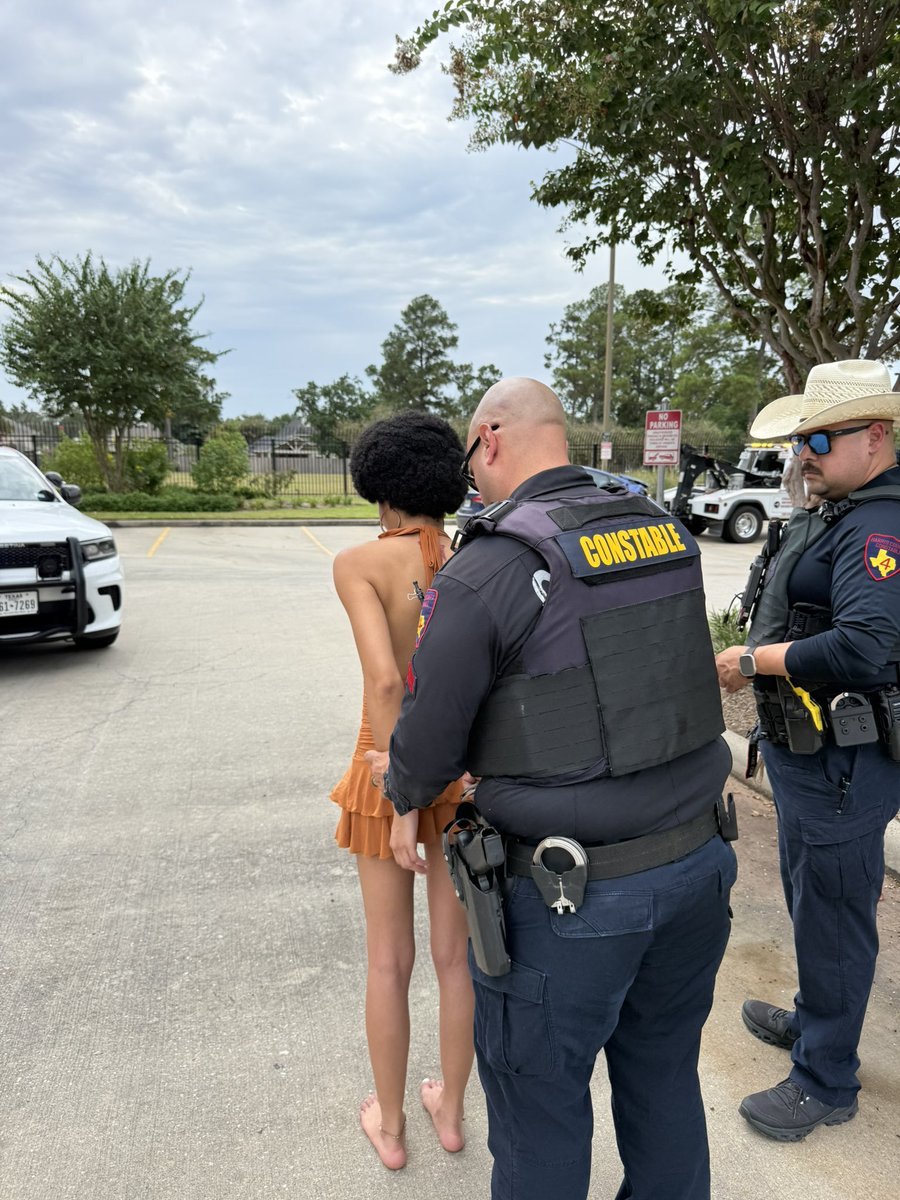 Two constable deputies in tactical vests detain a woman in an orange dress in a parking lot.
