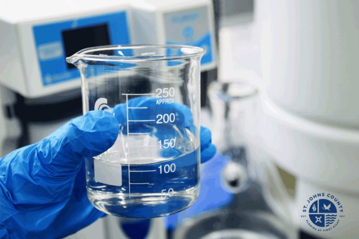Gloved hand holding a clear glass beaker with measurement markings in a lab.