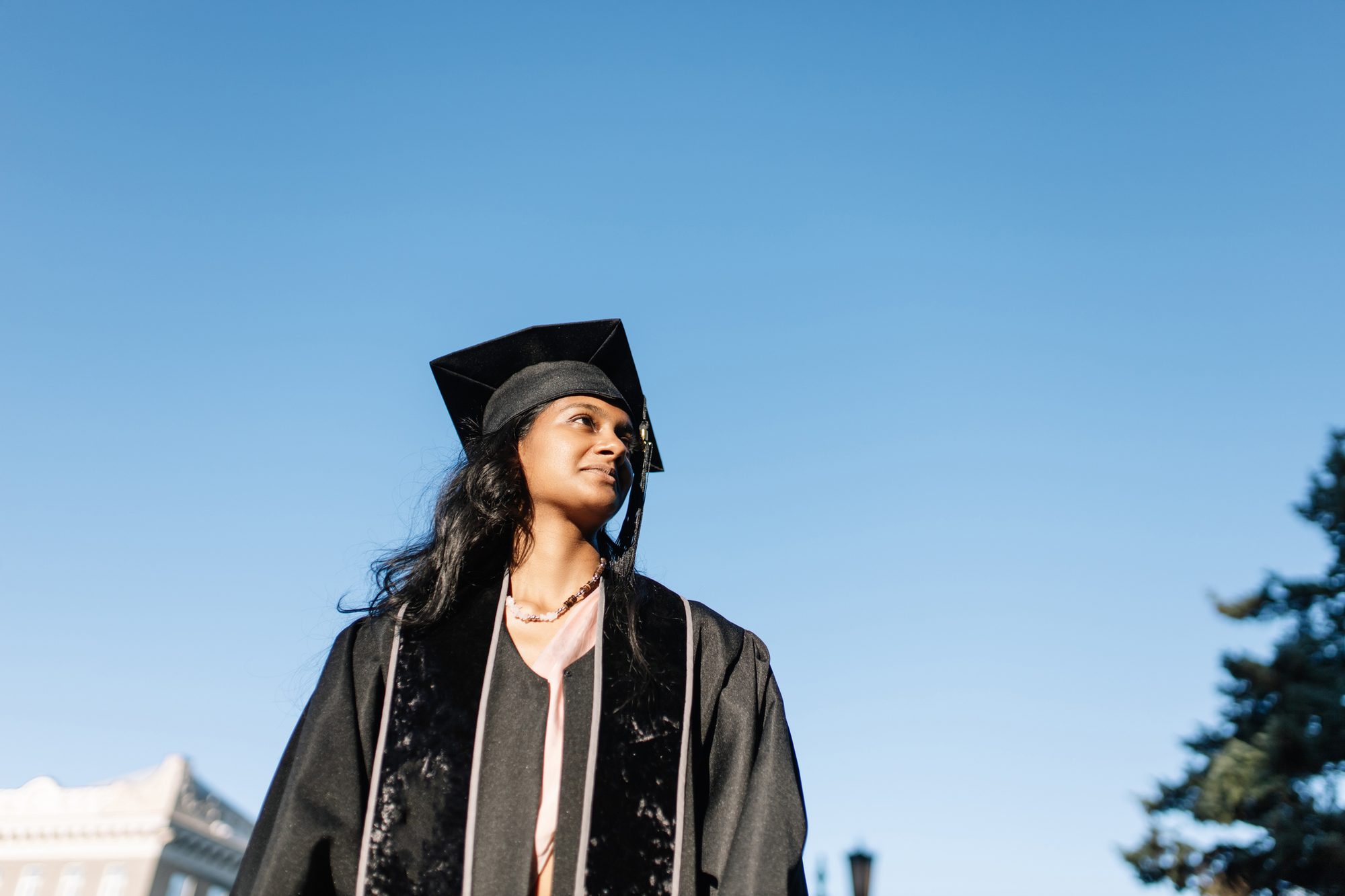 woman in graduation gown and mortarboard standing over clear sky, looking away with smile.