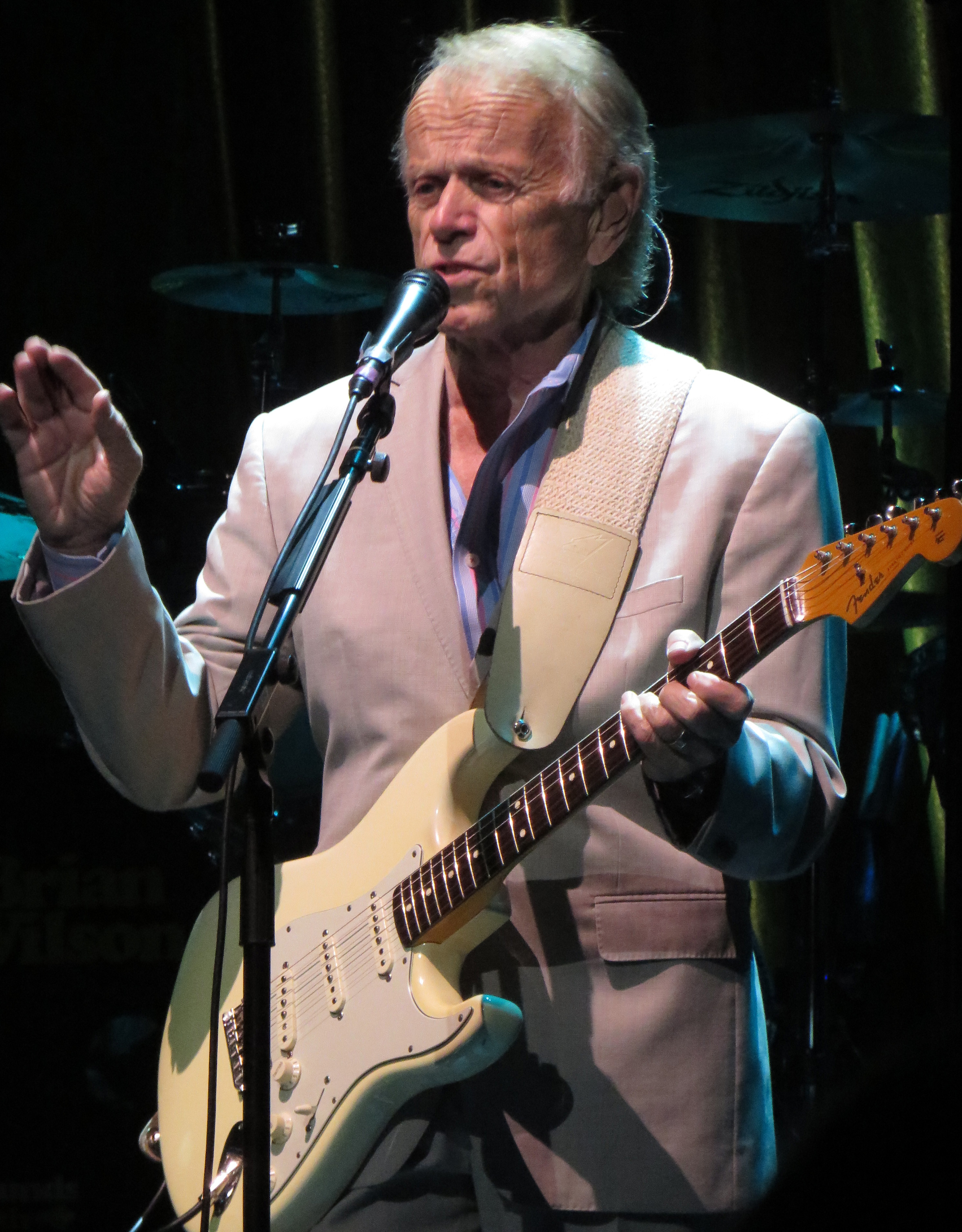 Older man in a light blazer plays electric guitar and sings into a microphone on a dimly lit stage.
