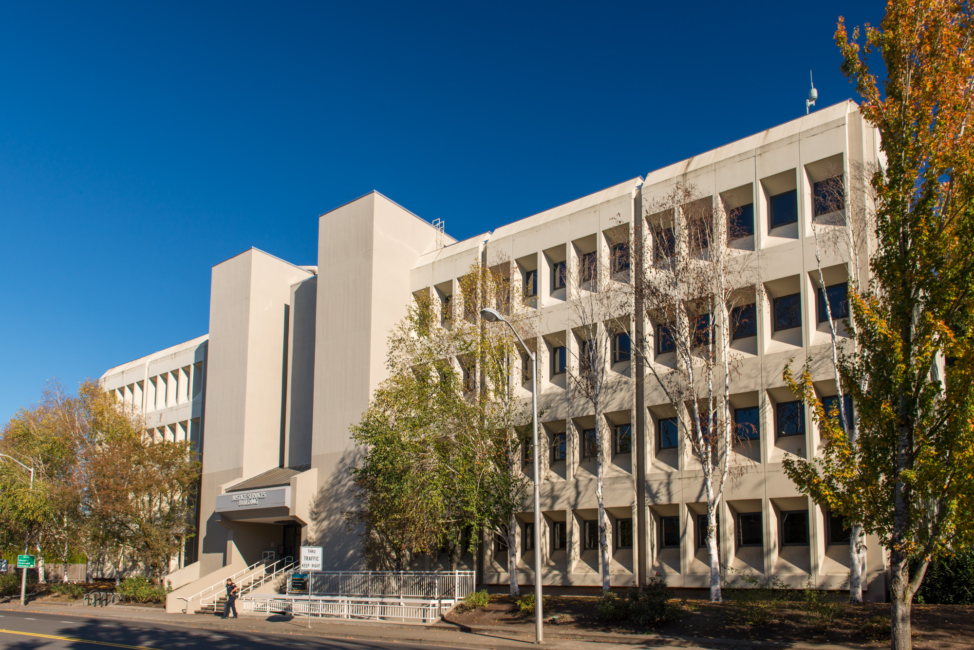 Multi-story modernist government building with beige exterior and autumn trees in front.