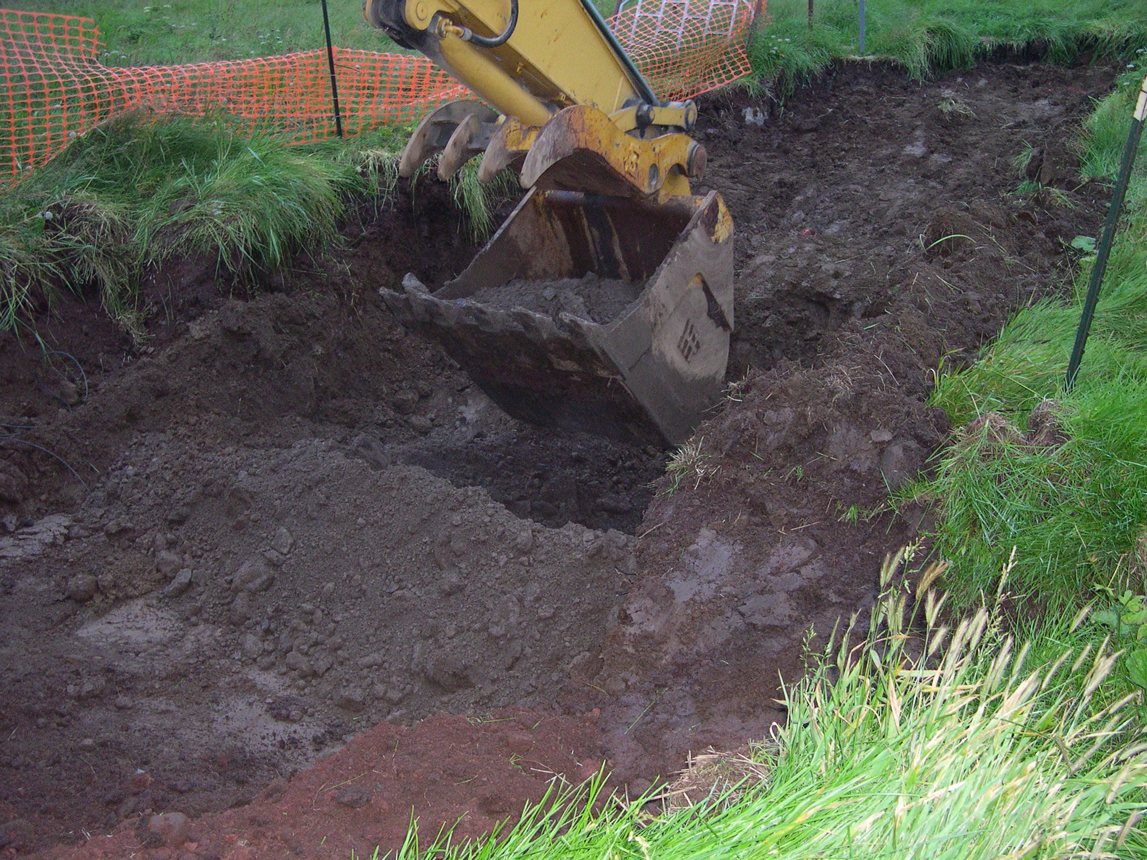 Yellow excavator bucket digging into dark muddy soil beside orange safety fencing.