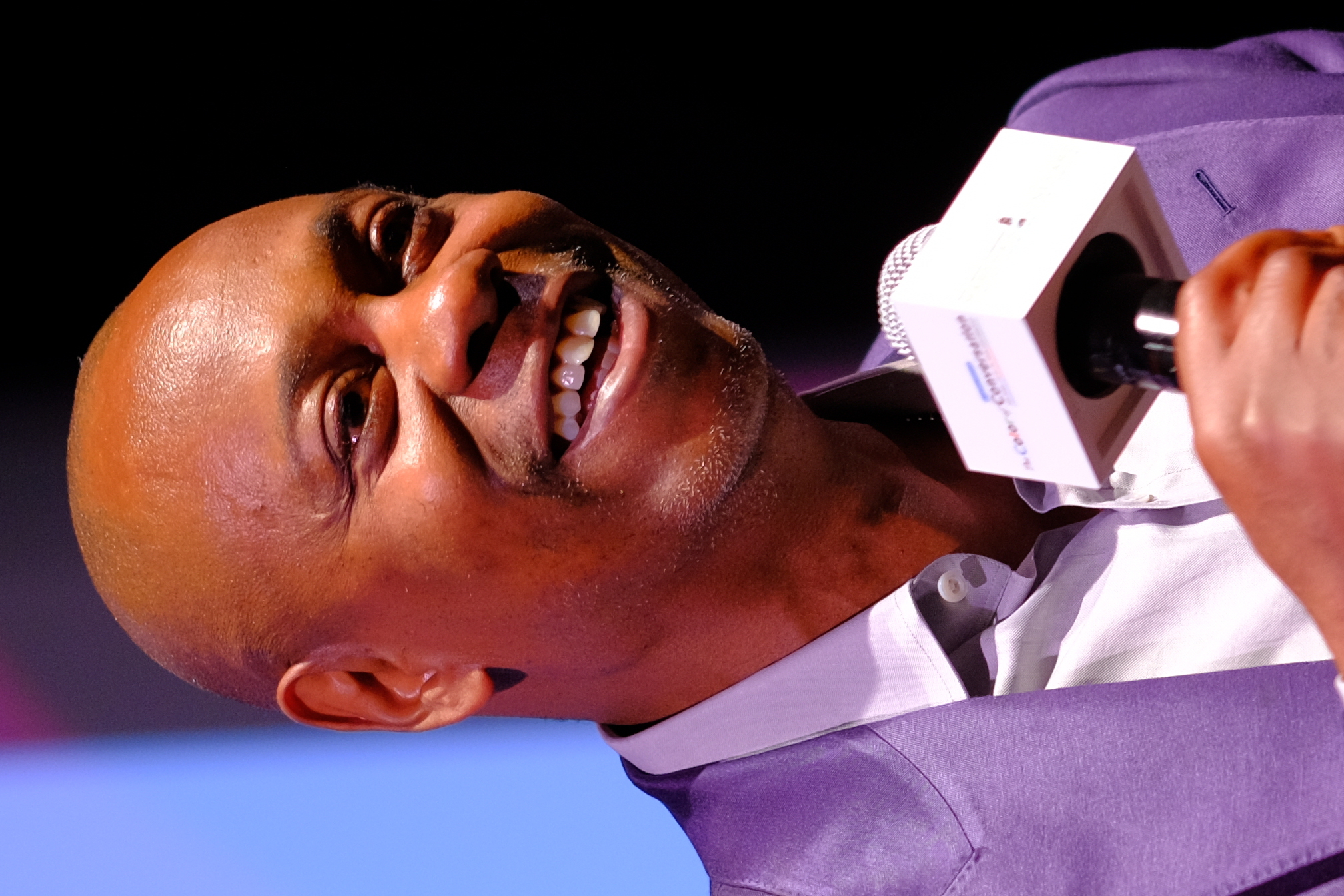 Man in a purple blazer smiling and holding a microphone at an indoor event