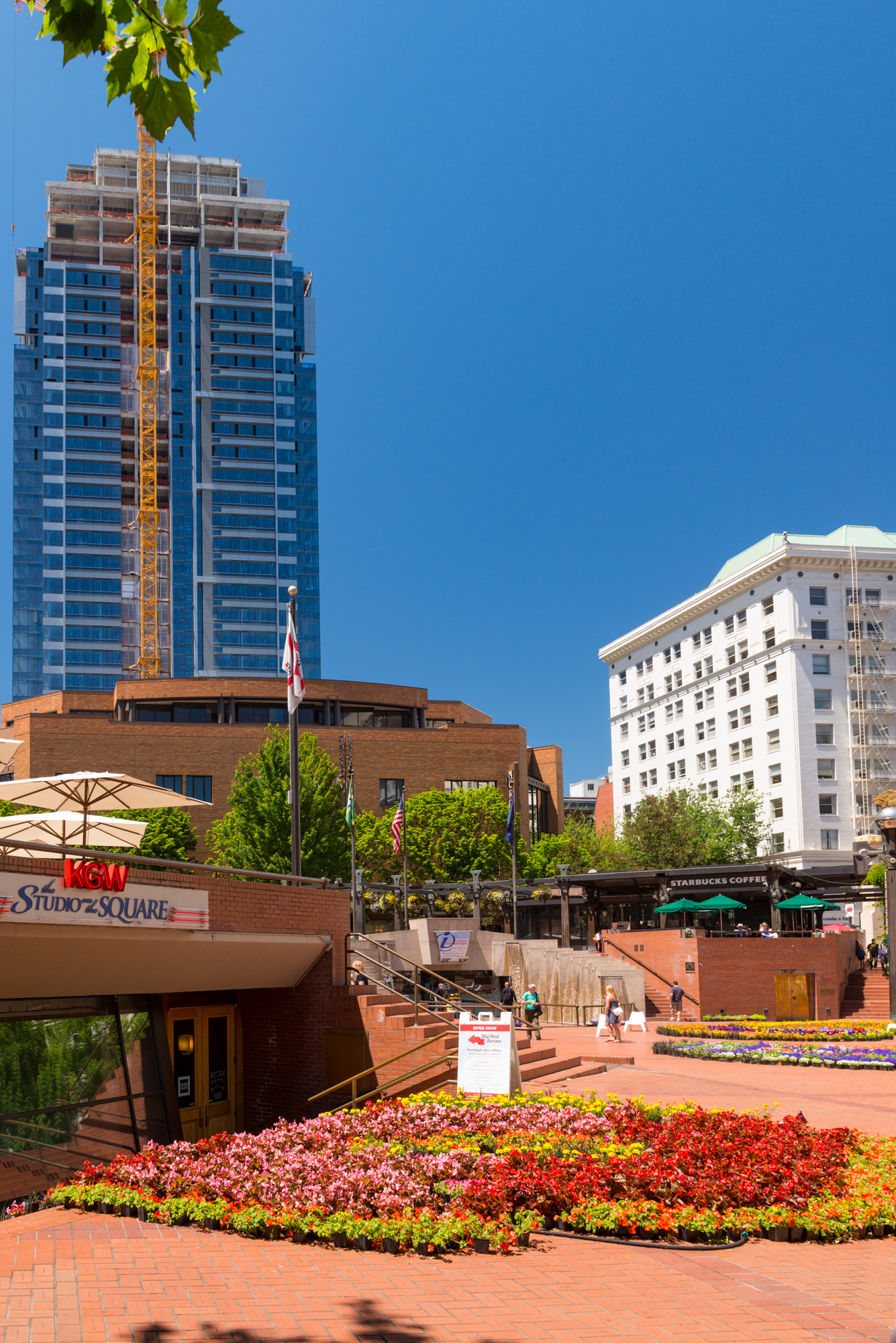 Sunny brick plaza with colorful flower beds, flags, and a tall glass building behind.