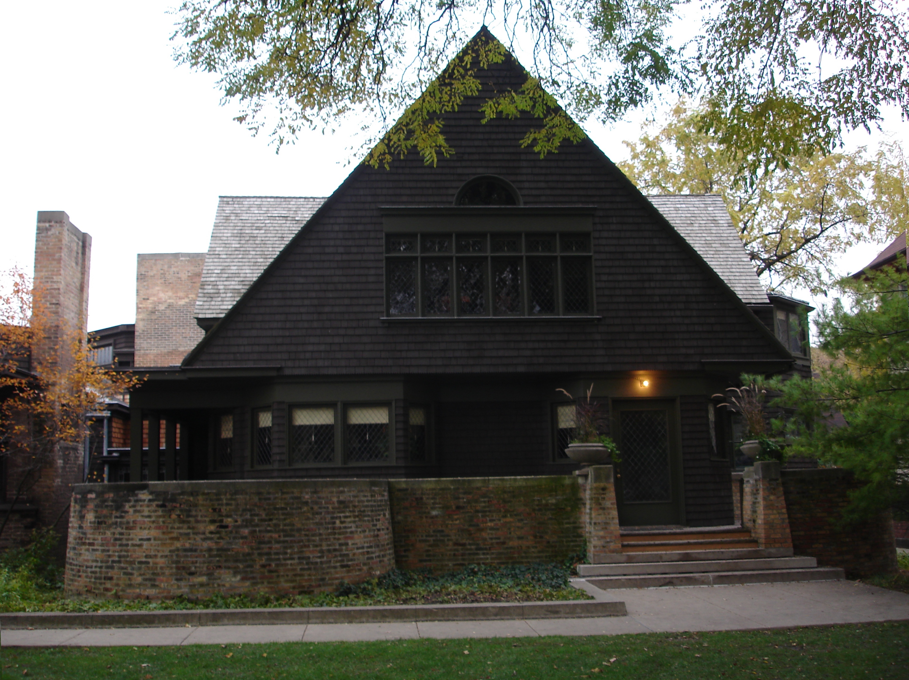 Dark brown shingle house with steep triangular gable and low brick wall.