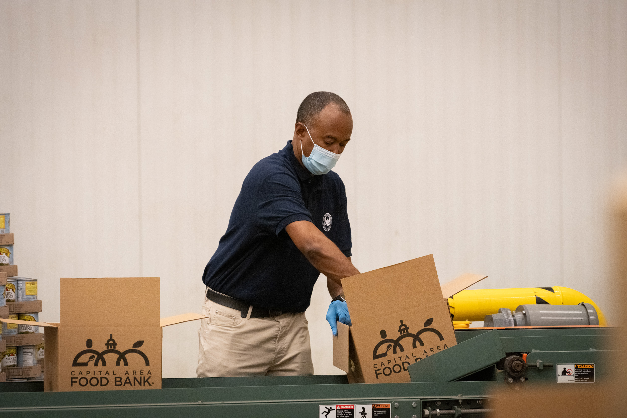 Masked worker in gloves handles labeled food bank boxes on a conveyor belt.