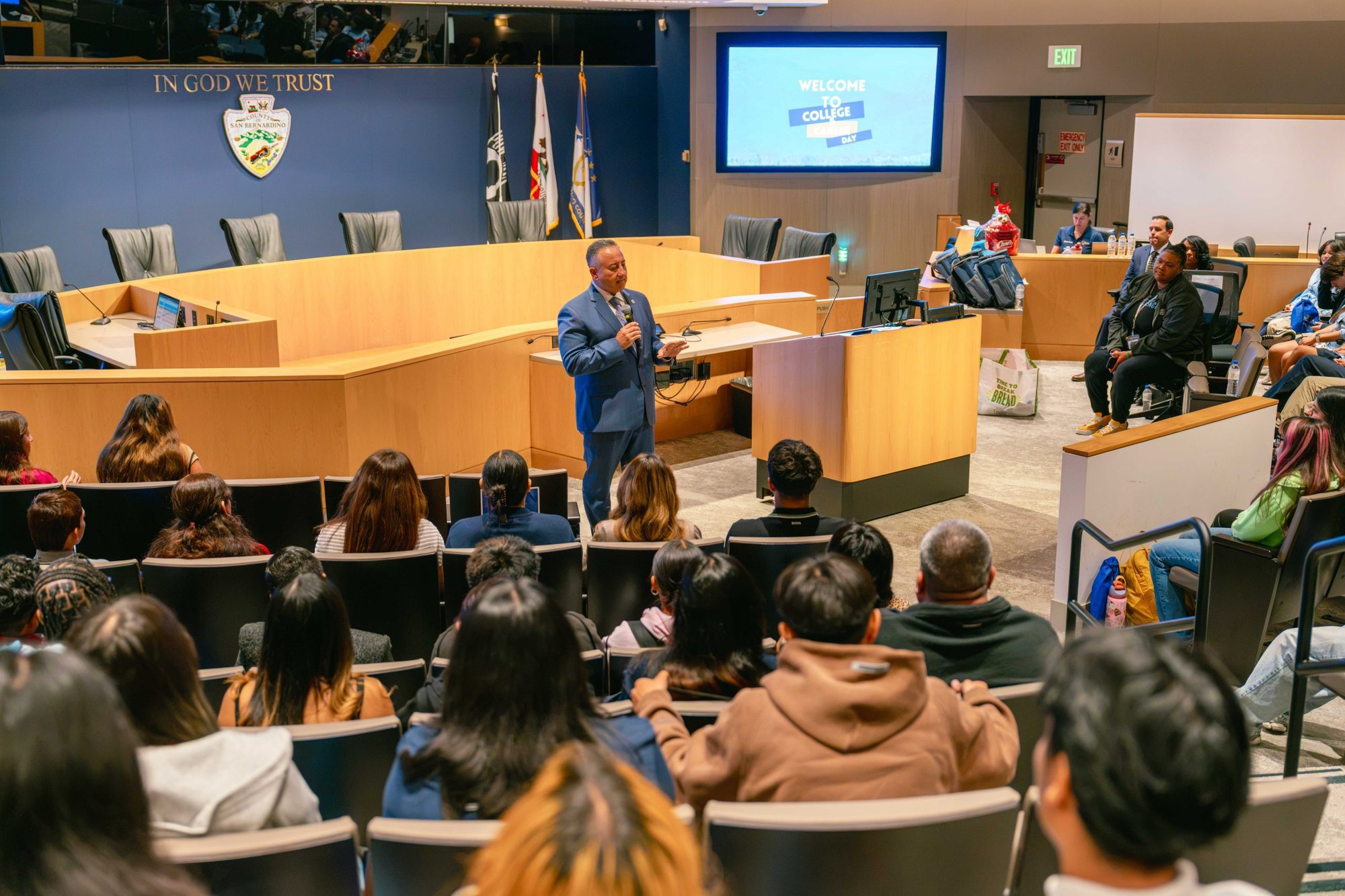 A man in a blue suit speaks at a podium before rows of seated students in a government chamber.