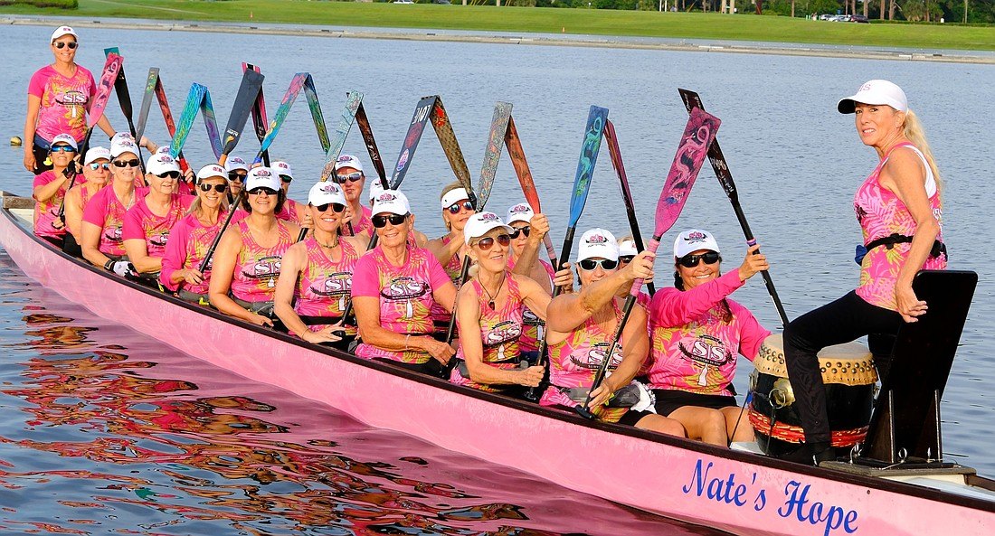 Women in matching pink shirts paddle a pink dragon boat on calm water.