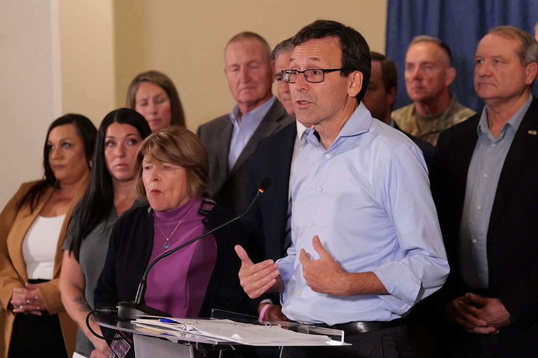 A man in a light blue shirt speaks at a podium before a group of officials at an indoor press conference.