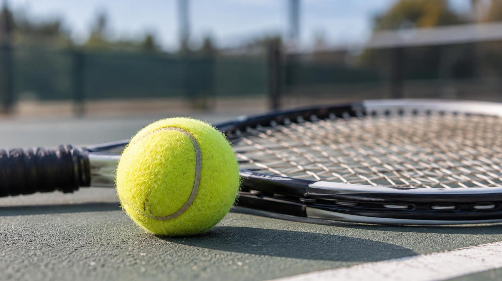 A yellow tennis ball resting on a hard court surface next to a tennis racket.