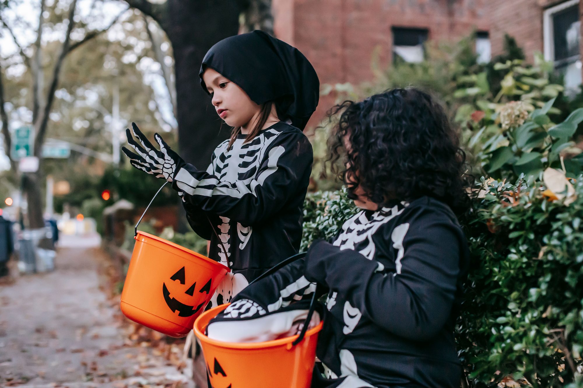 Serious adorable girls wearing skeleton costumes preparing for trick or treat with candy orange buckets painted like jack o lantern in daylight