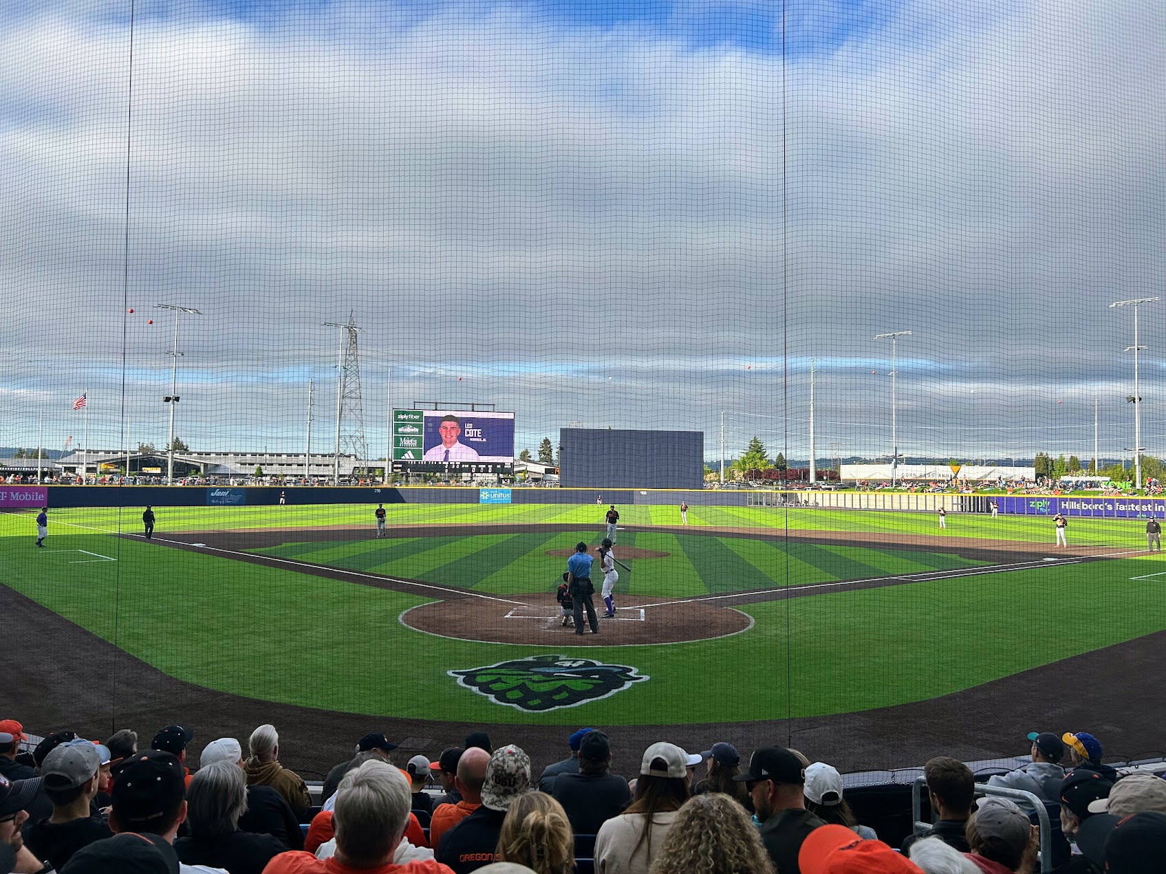 Wide view from behind home plate at a baseball stadium showing the field, scoreboard, and fans.