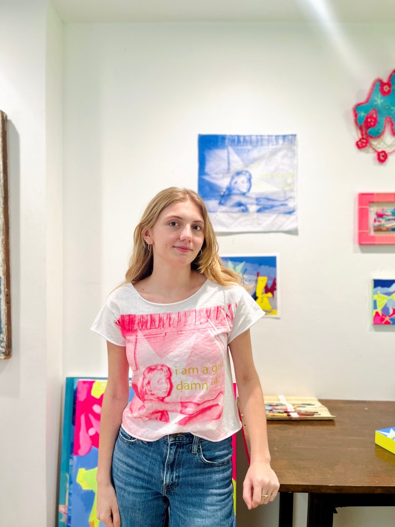 Young woman with blonde hair in a pink graphic t-shirt standing in an art studio with colorful paintings on the walls.