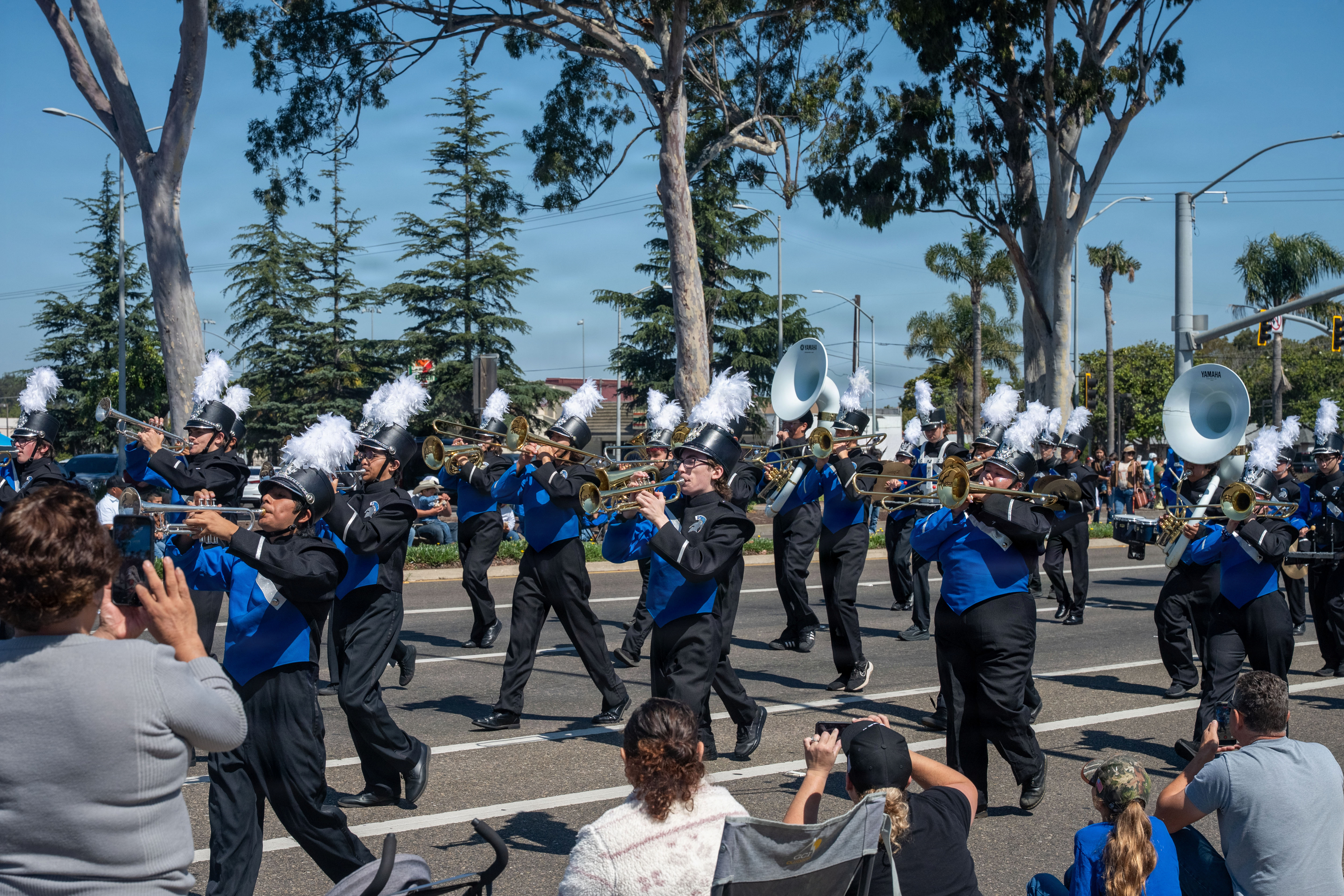 Marching band in blue and black uniforms playing brass instruments along a parade route.
