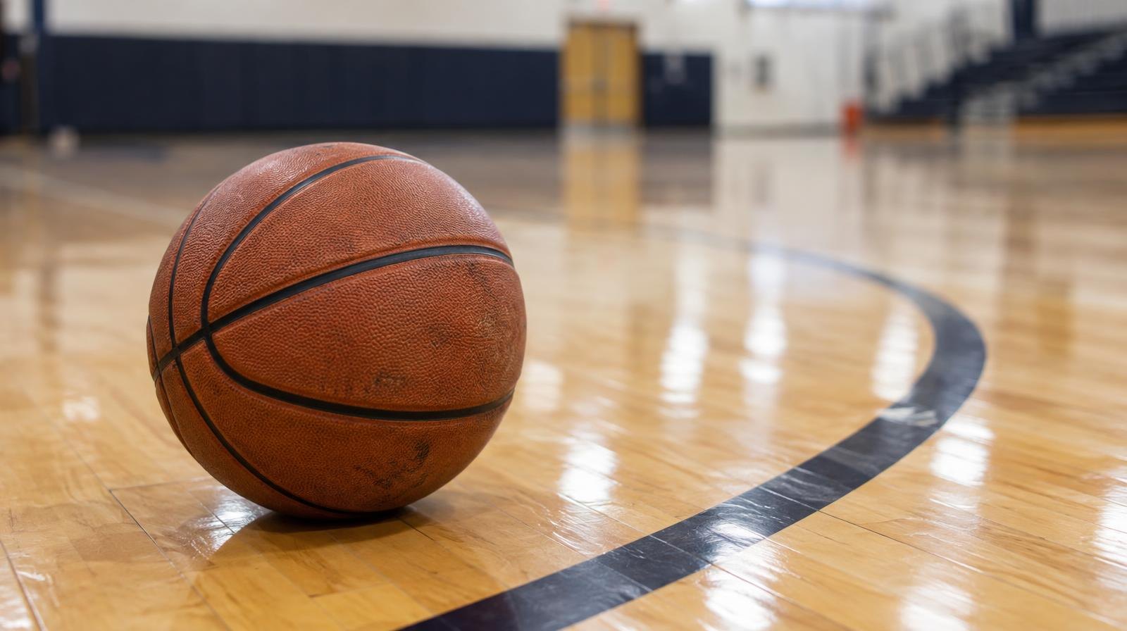A basketball resting on a hardwood floor beside a painted three-point arc.