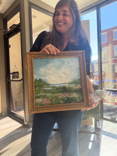 A woman holds a framed landscape painting inside a sunlit retail shop.