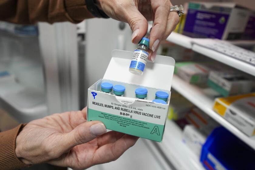 Hands holding a box of MMR vaccine vials with one vial lifted from the container.
