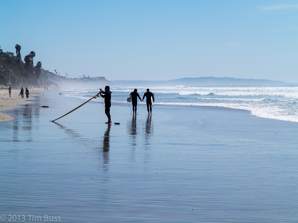 Three silhouetted figures walk along a wet sandy beach with coastal bluffs behind them.