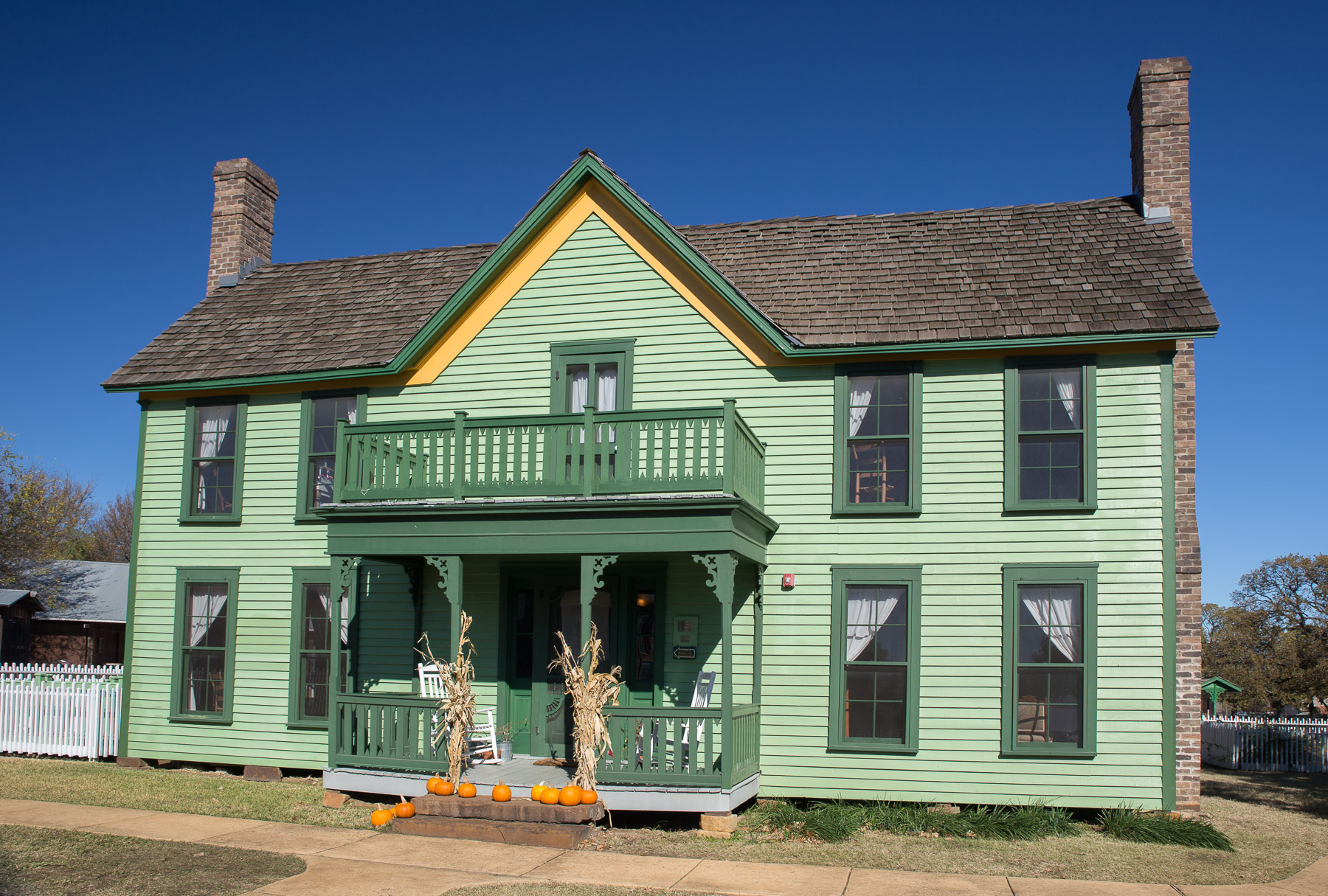 Two-story green farmhouse with fall pumpkins and corn stalks on the front porch.