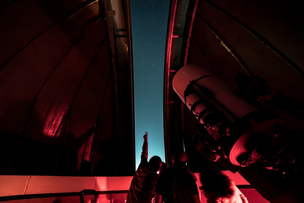 Silhouetted visitors stand inside a red-lit observatory dome, a large telescope pointing toward a starry sky.