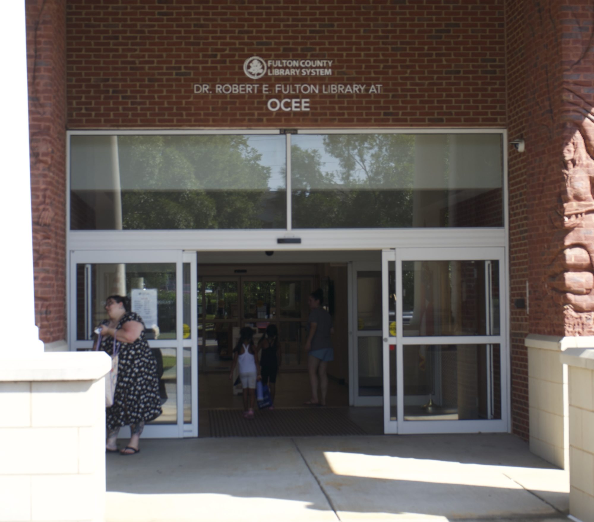 People entering and exiting a brick public library through glass doors on a sunny day.