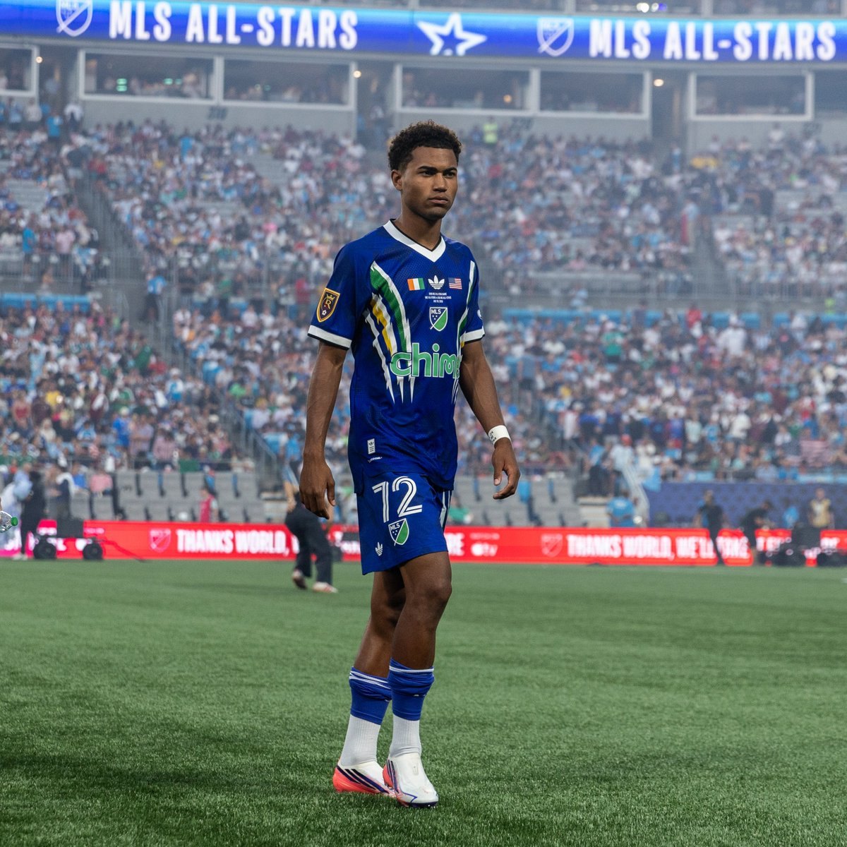 Soccer player in blue MLS All-Stars jersey, number 72, standing on a grass pitch before a packed stadium.