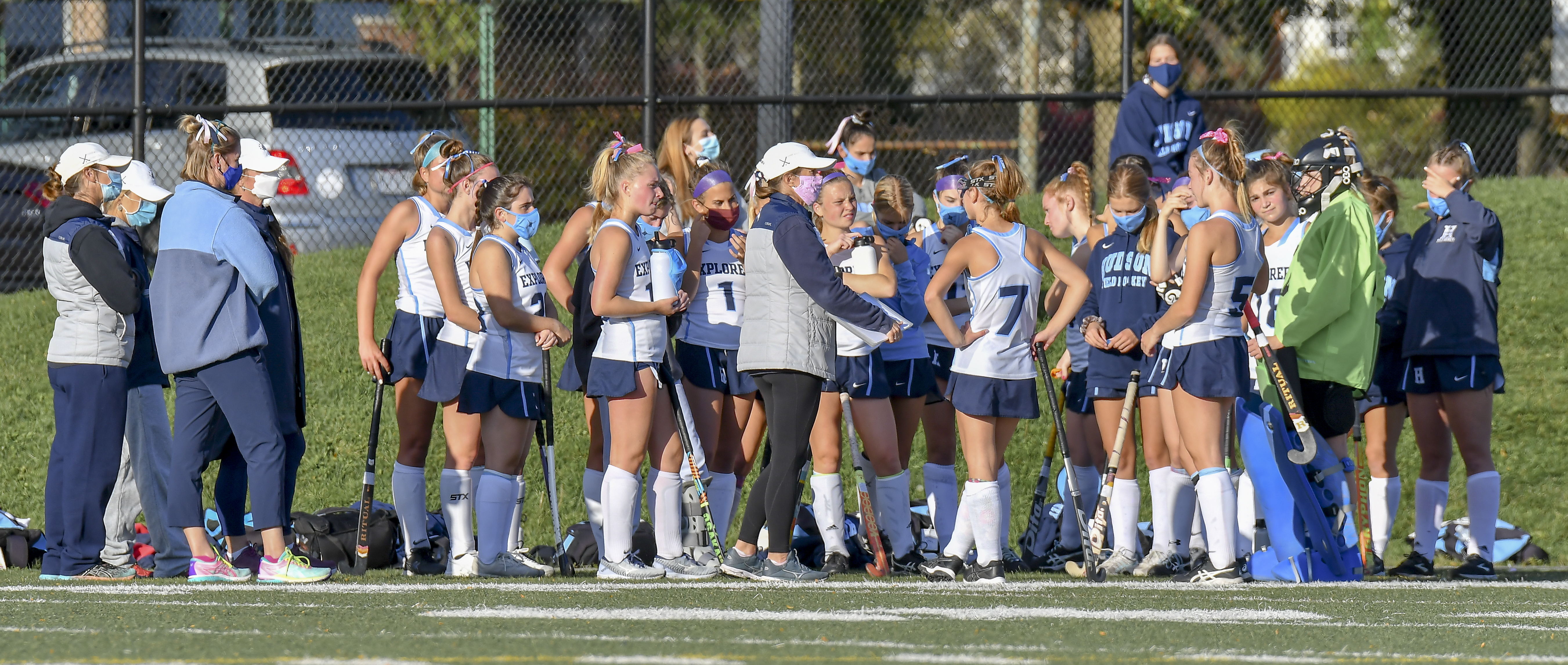 Female field hockey players in white and navy uniforms huddle on a turf field.