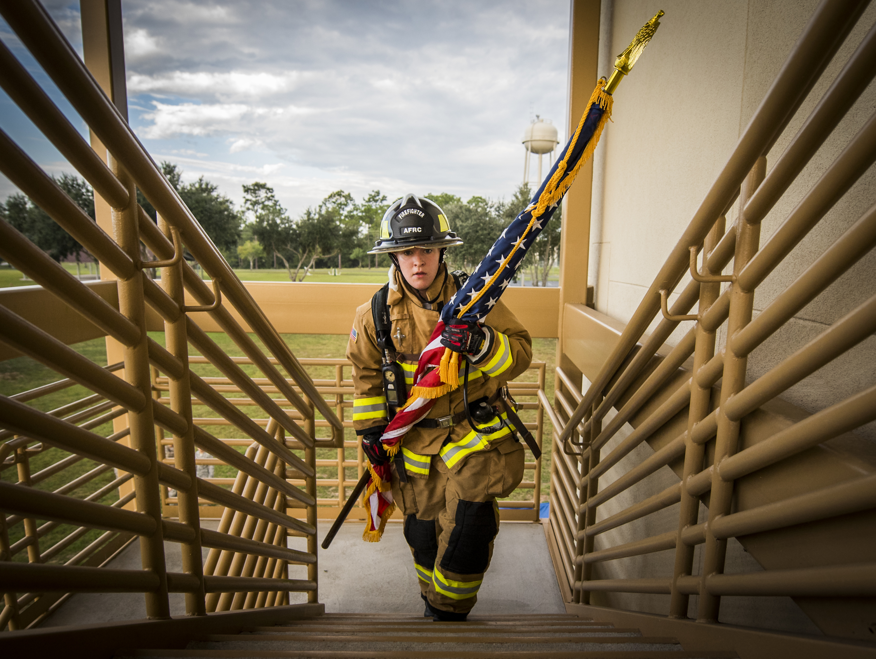 Firefighter in full gear and helmet carries an American flag up outdoor stairs.