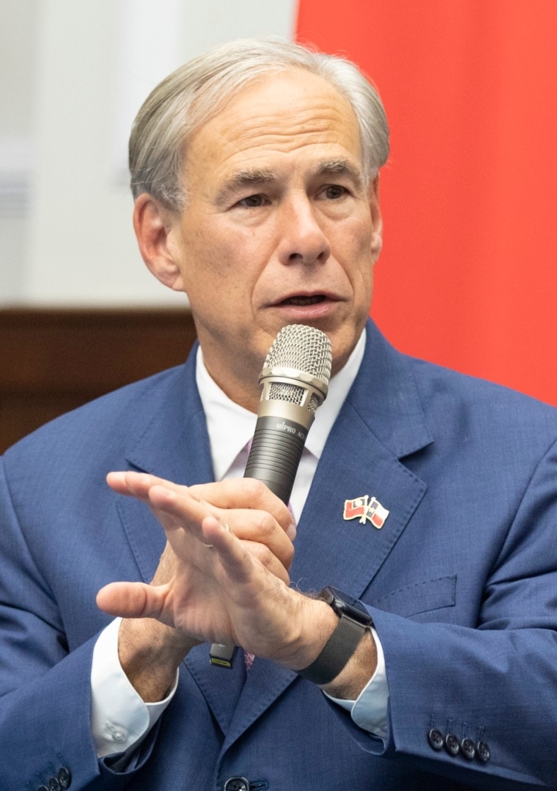 Man in blue suit speaking into a handheld microphone against a red backdrop.