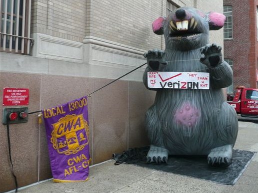 Giant inflatable protest rat beside a CWA AFL-CIO union banner on a city street.
