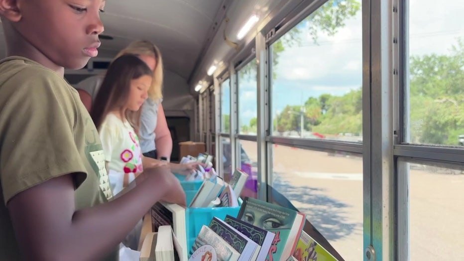 Children selecting books from shelves inside a converted bus with large windows.