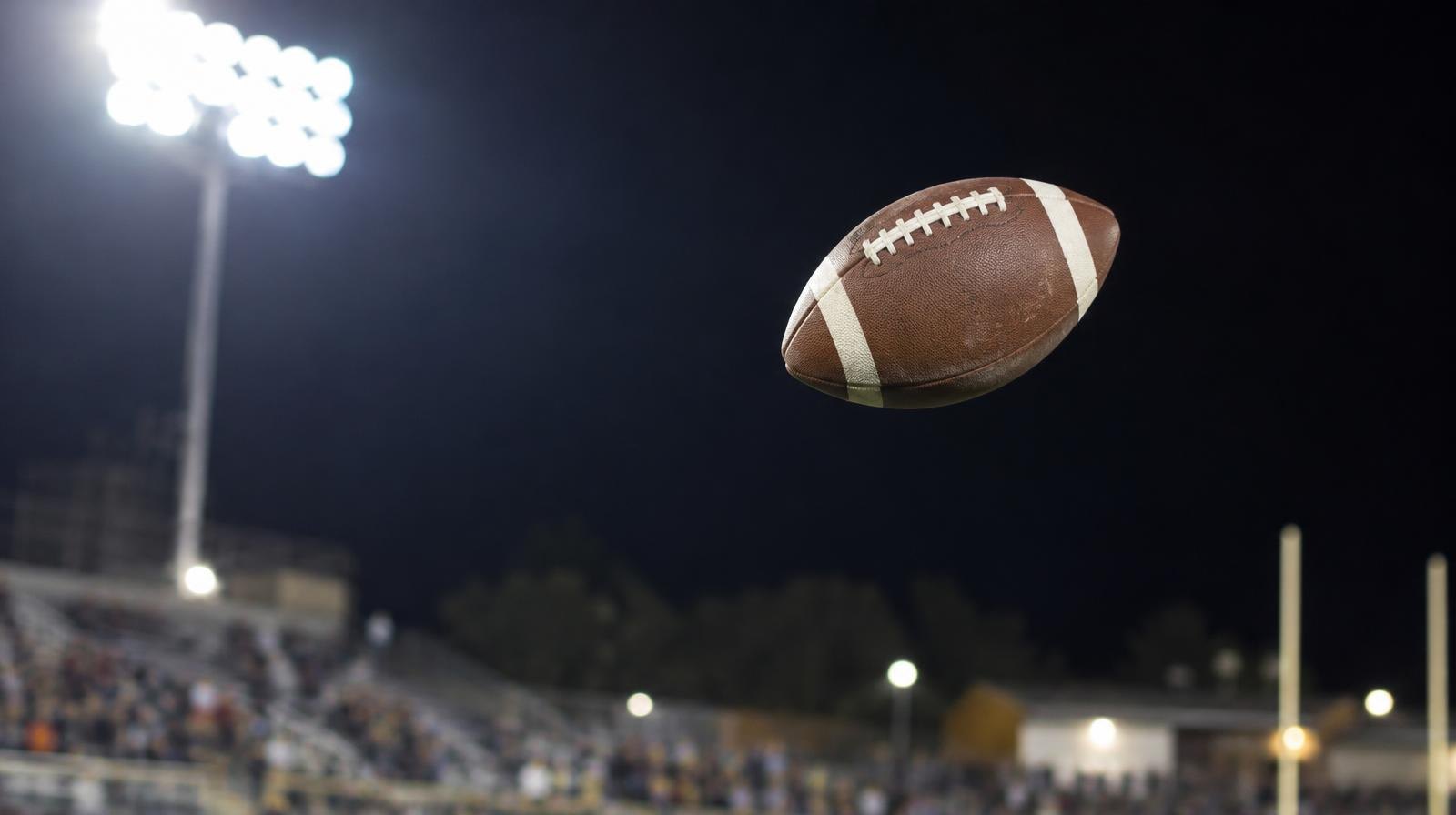 A football spiraling through the air under bright stadium lights against a dark night sky.