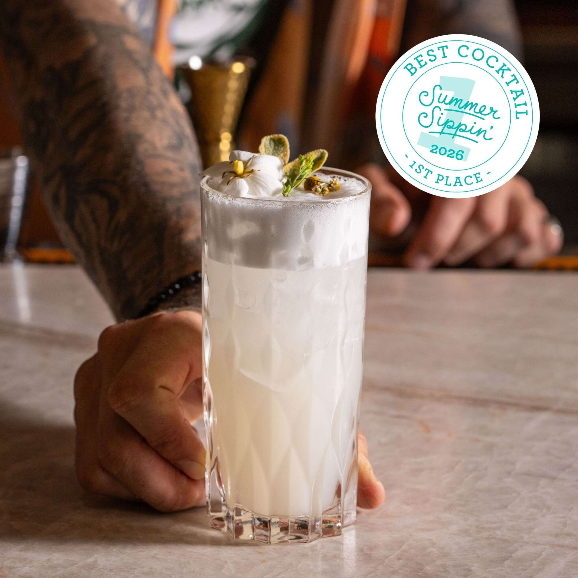 A frothy white cocktail in a tall faceted glass garnished with herbs and flowers, held by a tattooed hand.