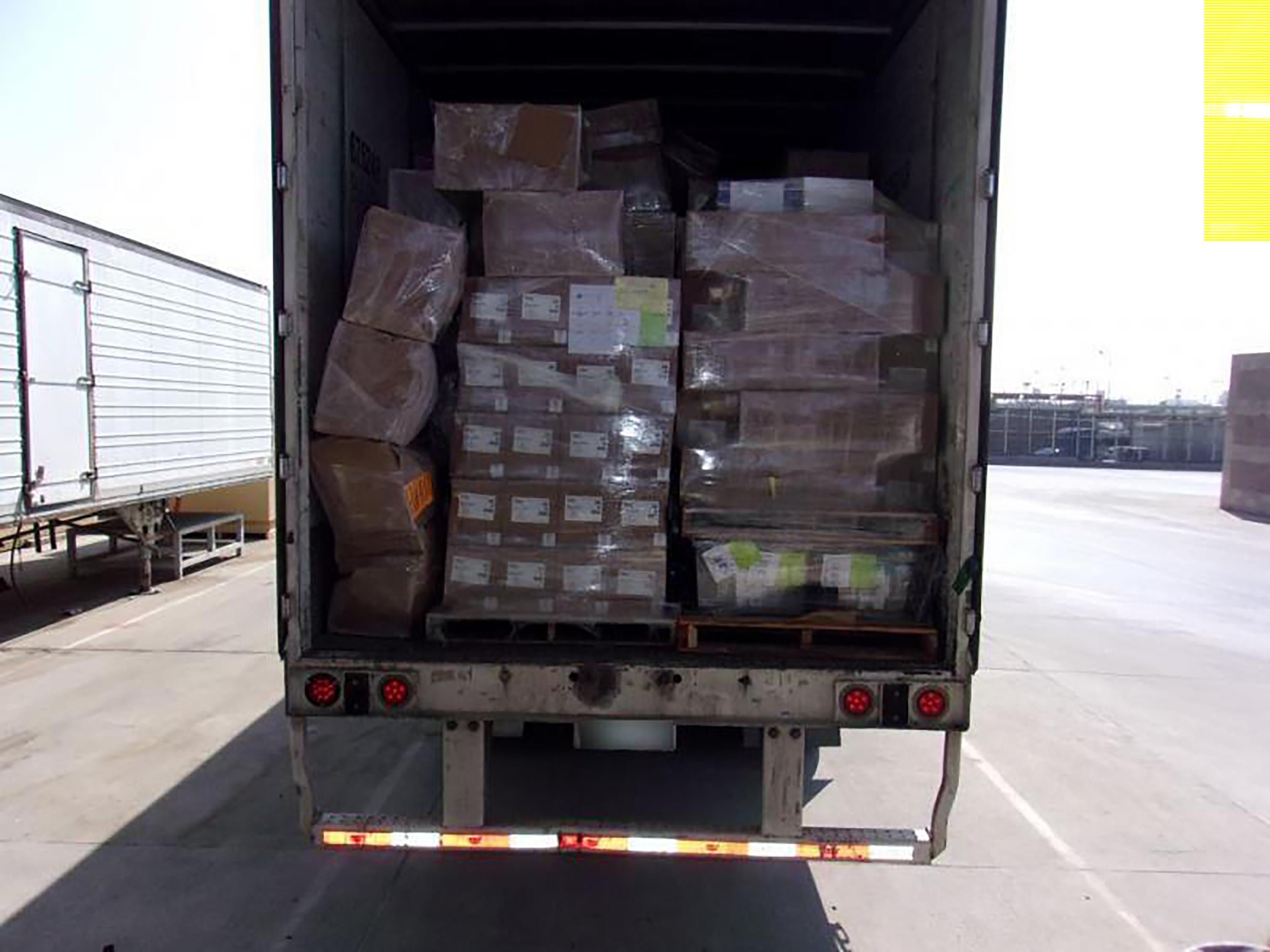 Open rear doors of a semi-trailer loaded with pallets of plastic-wrapped boxes at a cargo facility.