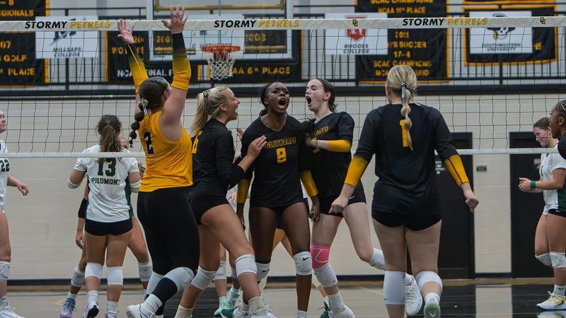 Women's volleyball players in black and gold uniforms celebrate on an indoor court.