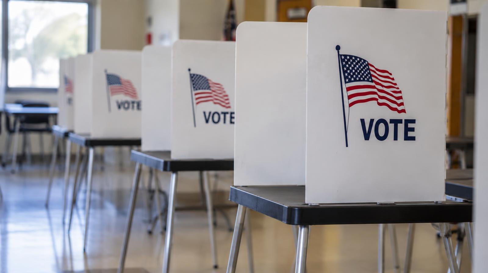 A row of empty voting booths with privacy screens in a brightly lit hall.