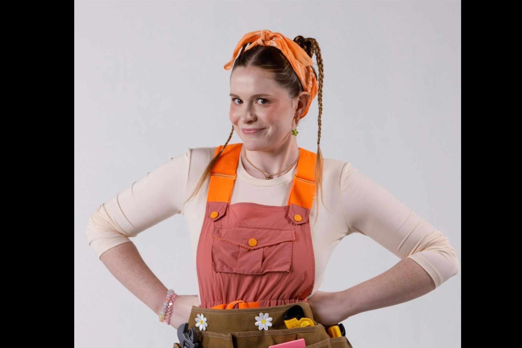 Young woman posing in pink plumber overalls with orange headband and daisy-decorated tool belt in a studio.