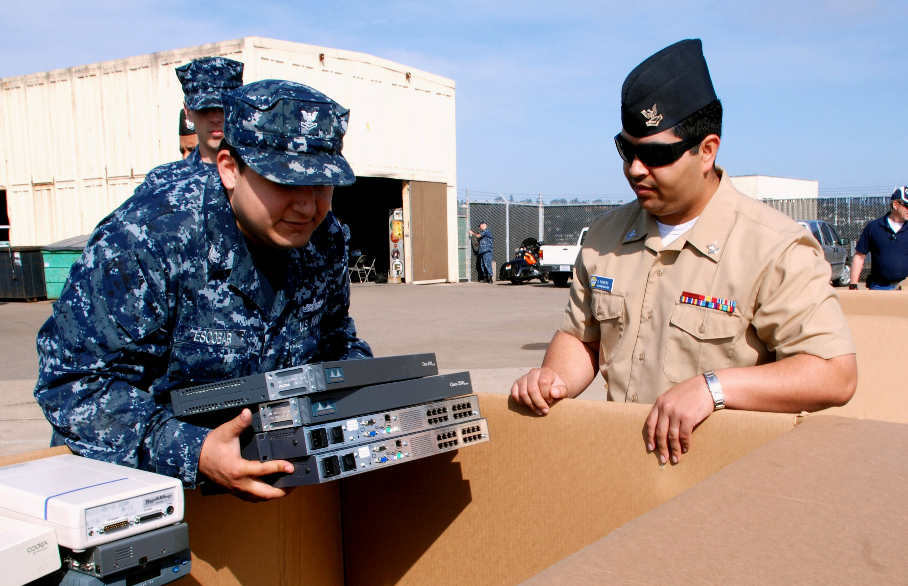 Two people in uniform carrying electronic equipment at an outdoor recycling event.