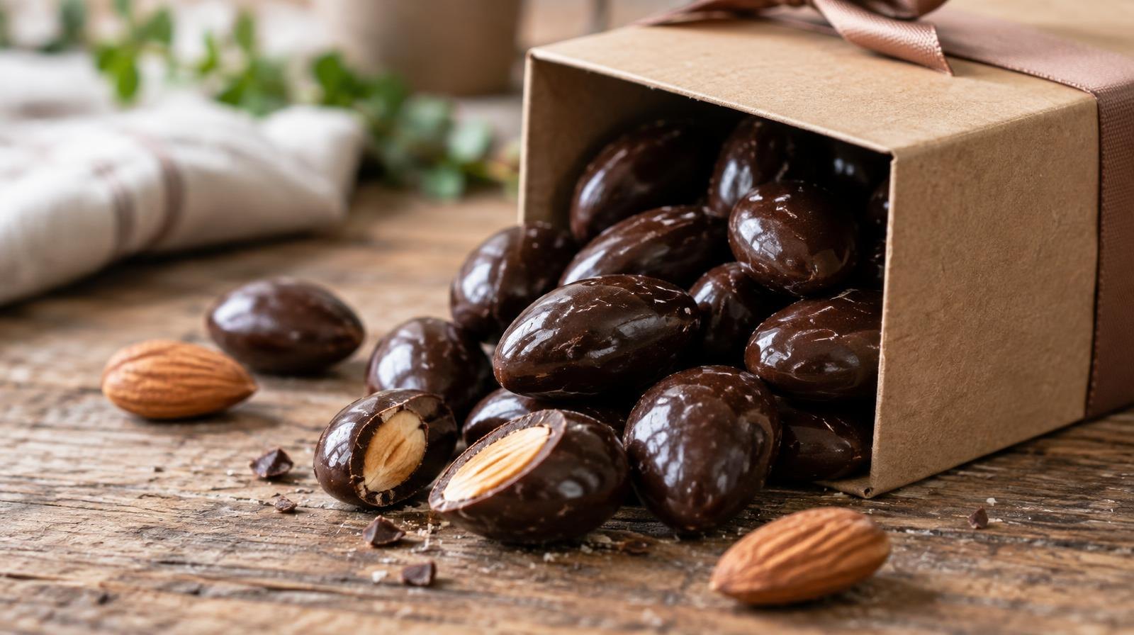 Dark chocolate-covered almonds spilling from a small kraft paper gift box on a wooden surface.