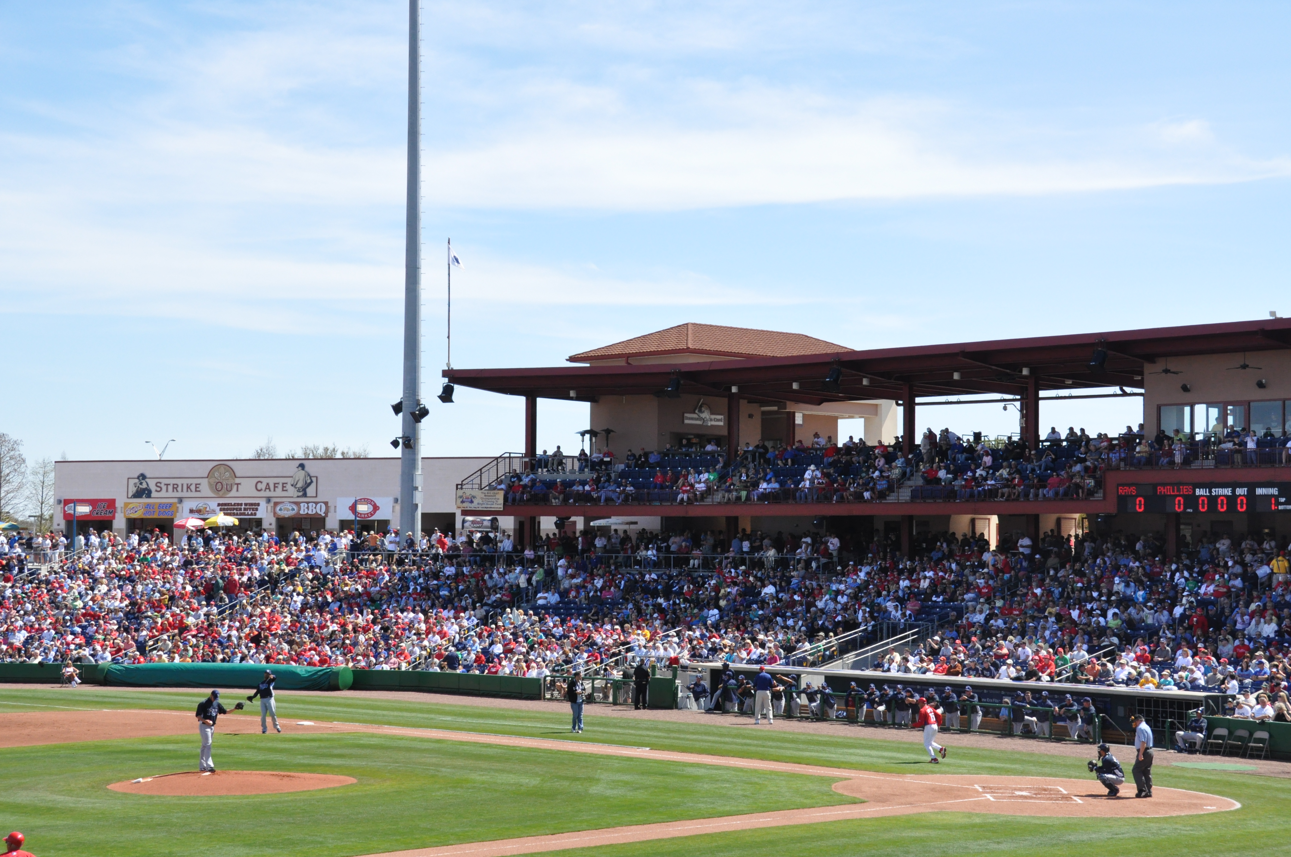 A packed minor league baseball stadium with players on the field during a sunny daytime game.