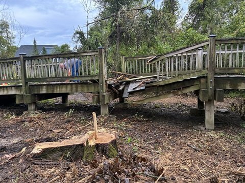 Damaged wooden pedestrian bridge with broken railings, debris, and a cut tree stump nearby.