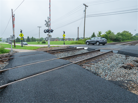 Railroad grade crossing with new dark rubberized surface, signal arms, and a car beyond the tracks.