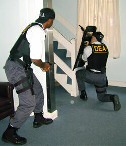 Two DEA agents in tactical vests conduct a staircase entry, one holding a ballistic shield.