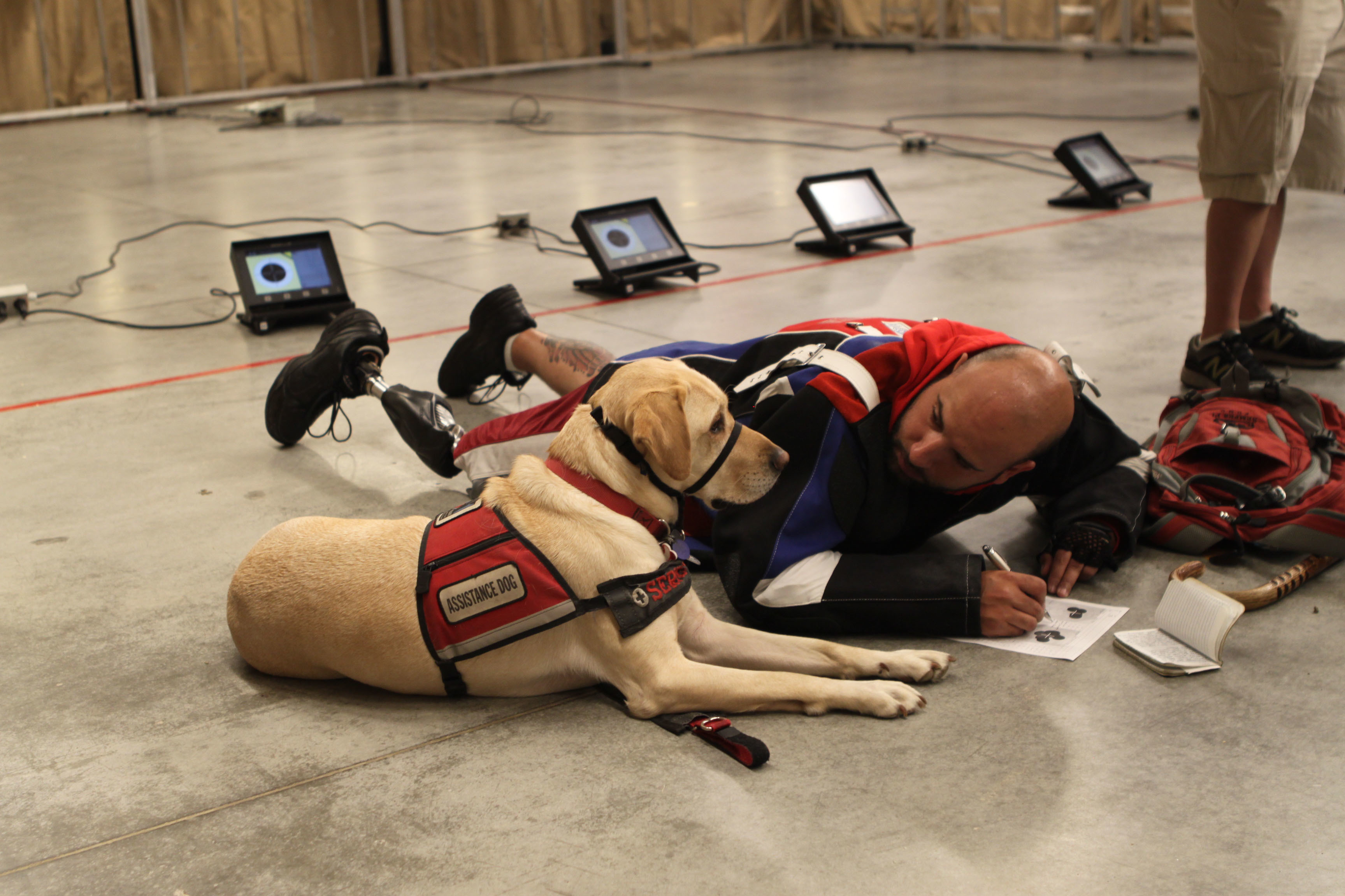 Yellow Labrador wearing a red service dog vest lies beside a man on a concrete floor.